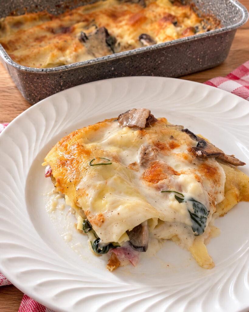 Porzione di lasagne ai funghi cardoncelli e salame spagnolo con la teglia sullo sfondo