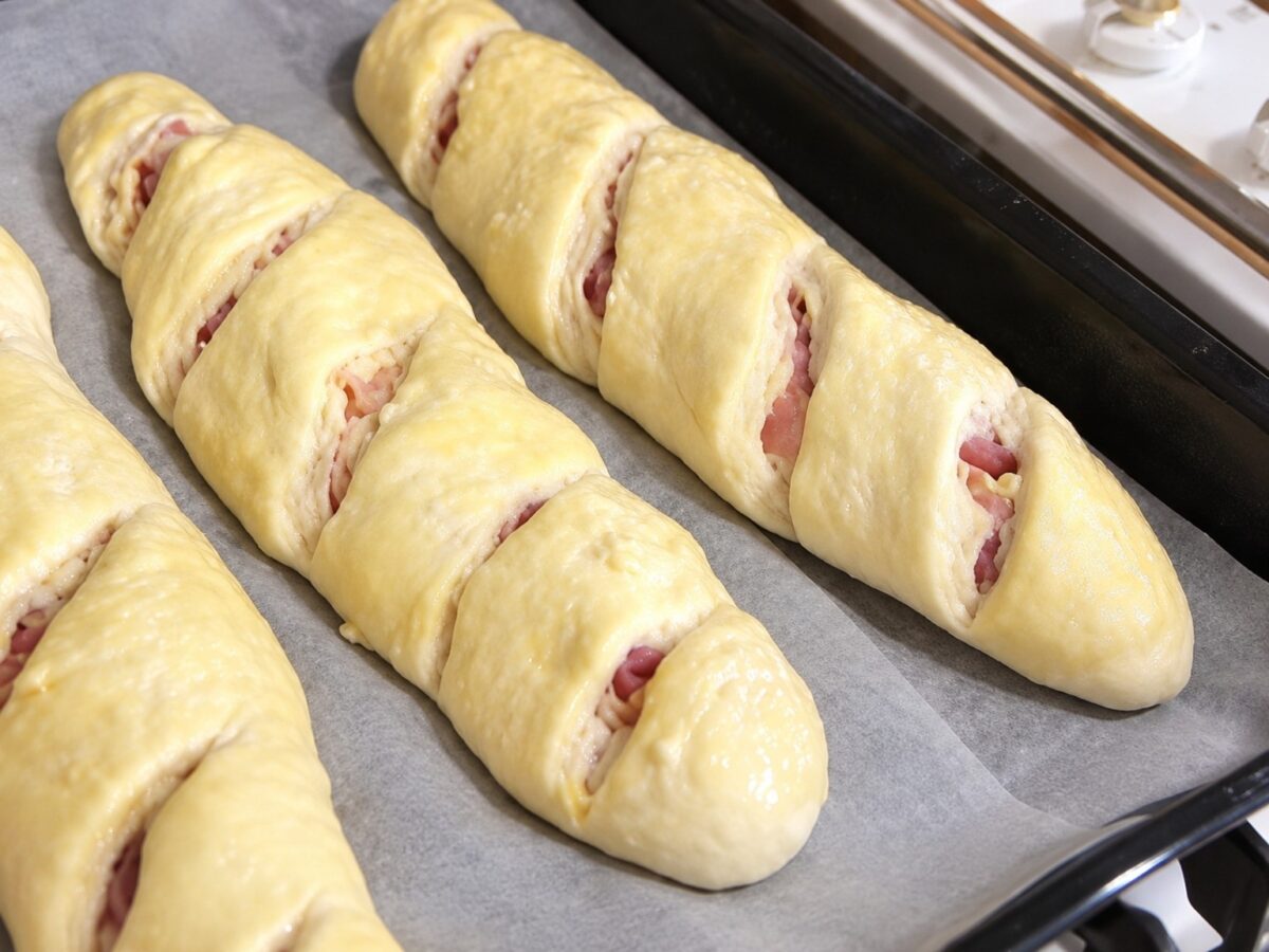 Baguettes farcite con prosciutto cotto, spennellate e incise, disposte sulla teglia prima della cottura